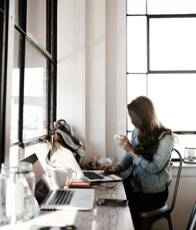 A young woman working on her laptop, sipping coffee in a bright café setting.