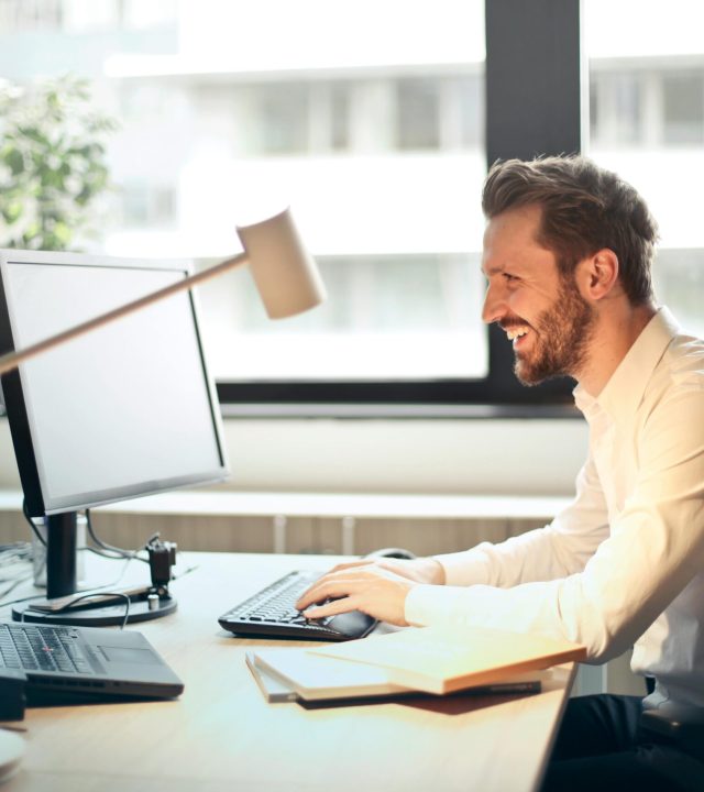 A man smiling while working at an office desk with a computer and natural daylight streaming in through large windows.